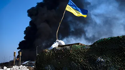 2022年3月25日,基輔郊區的一面烏克蘭國旗,背後則是因為俄軍空襲冒出的大量黑煙。(美聯社)