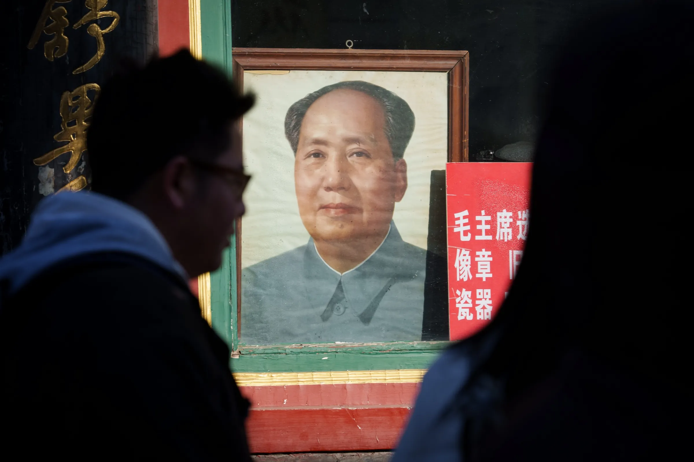 2026 年 1 月 12 日，北京街頭一家店舖外懸掛著前中國領導人毛澤東的畫像，行人匆匆走過。（美聯社）