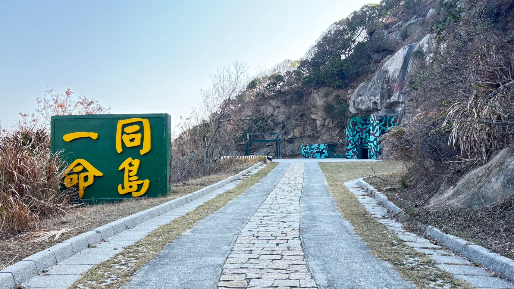 同島一命入口景觀，道路沿著岩壁延伸，軍事偽裝色建物與自然地景交錯，呈現戰地島嶼獨特風貌。(照片/鄧亦涵攝)