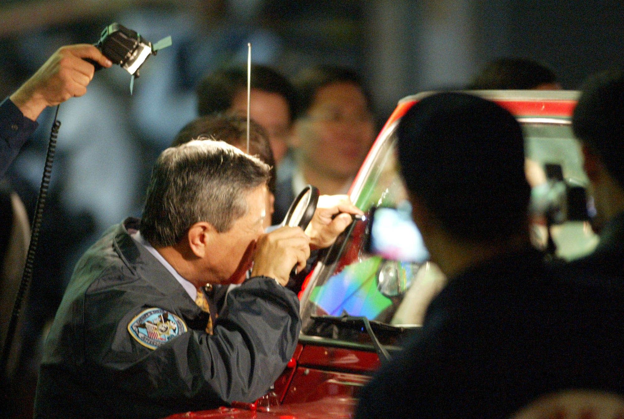 2004年4月9日，李昌鈺博士在台南檢視陳水扁總統遇刺，並用放大鏡檢視擋風玻璃上的彈孔。陳總統遇刺未遂兩週多後死亡。（美聯社）