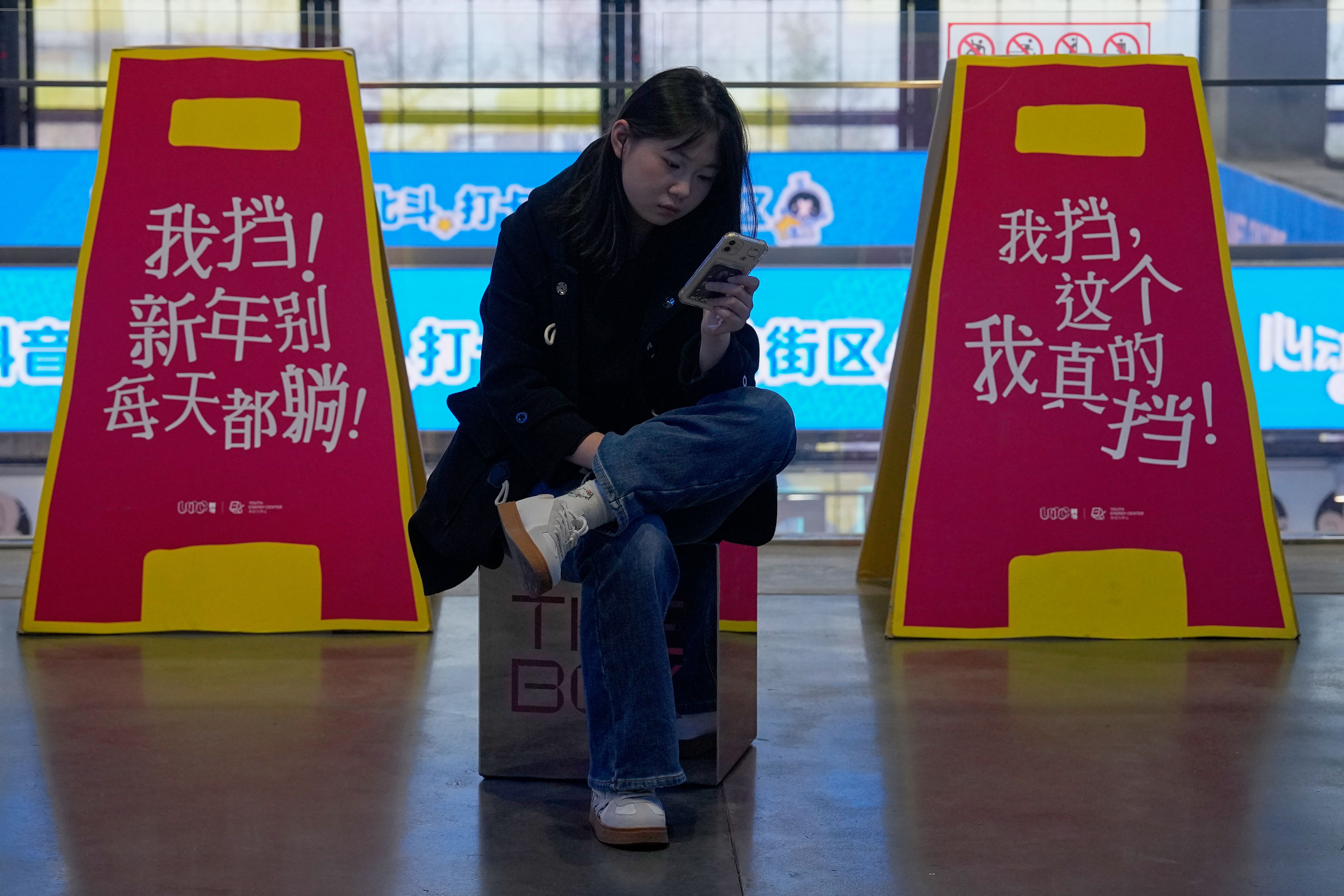 北京一名年輕女孩在購物中心划手機，後方是宣傳遠離負面生活態度的標語。（美聯社）