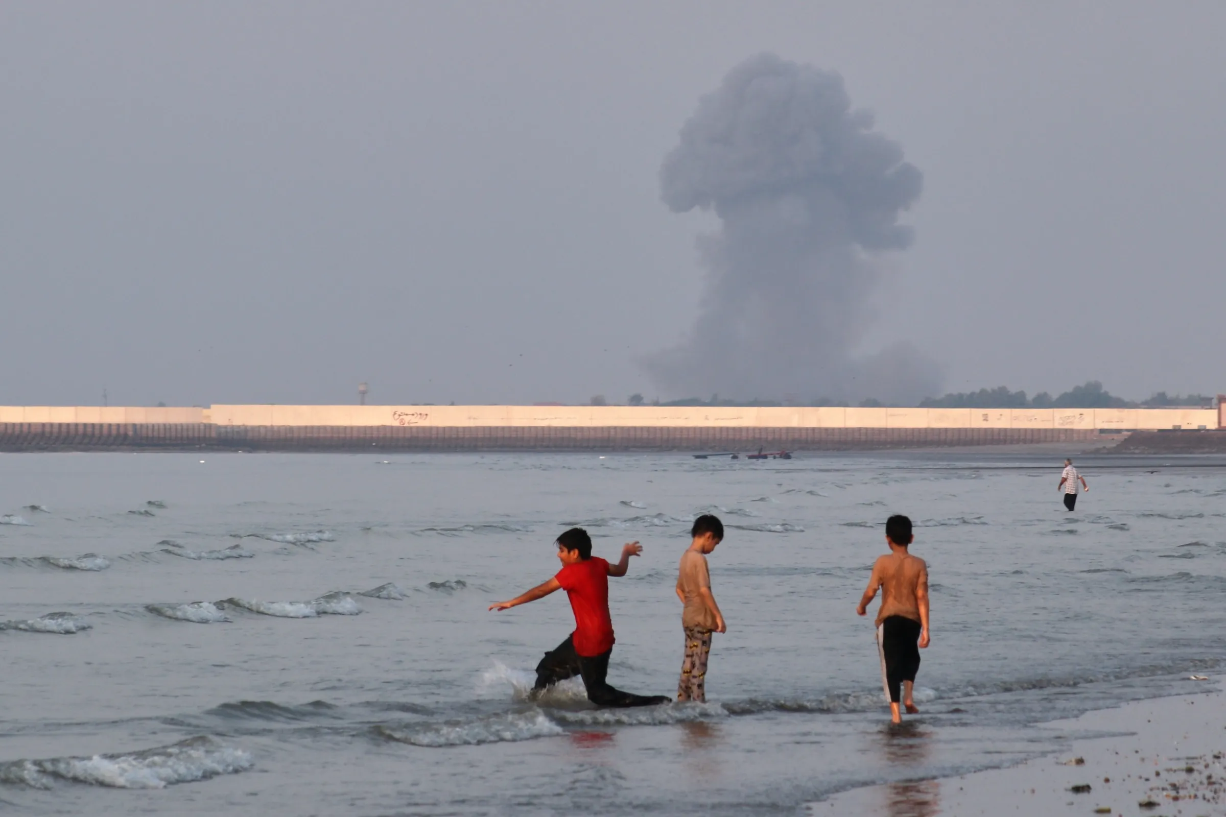2026年7月12日，伊朗小朋友在海邊玩耍，遠處就是荷莫茲海峽，還有爆炸引發的大量黑煙。（美聯社）