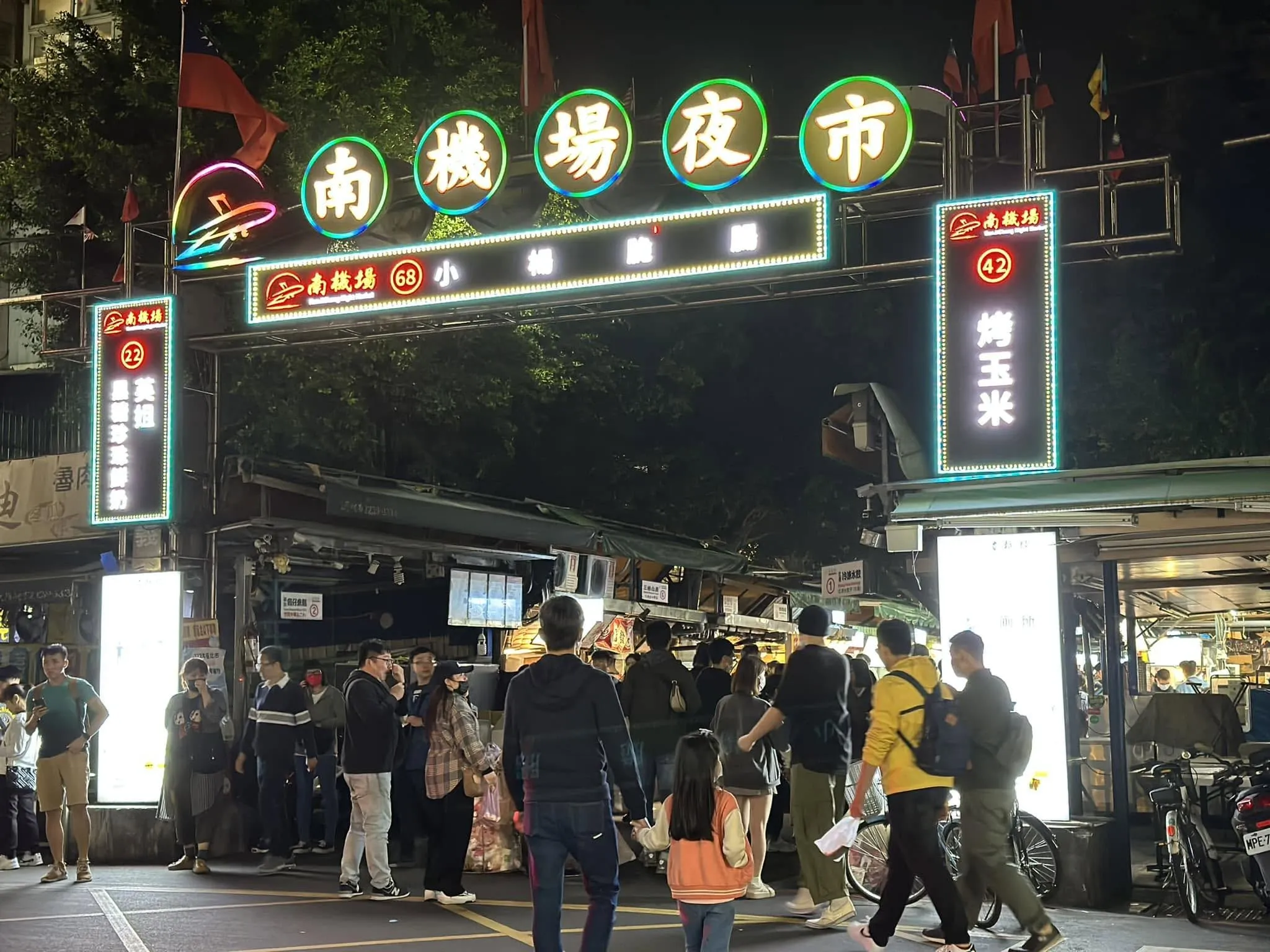 南機場夜市圓形綠光霓虹牌樓入口夜景。標示牌列出烤玉米與麻油雞等招牌攤位，入口處吸引大批饕客走訪，呈現台北市密度極高且無遊戲攤位之純美食天堂特色。（圖／取自FB＠南機場夜市）