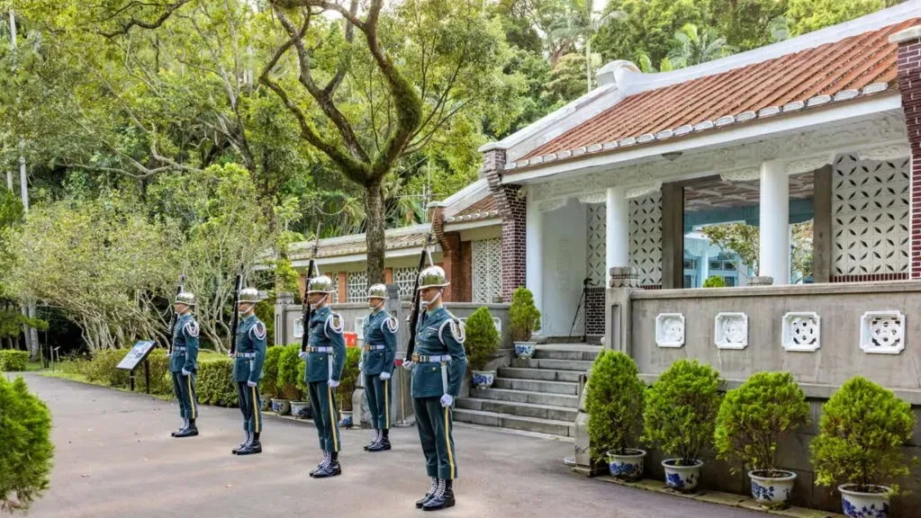 桃園景點慈湖陵寢儀隊交接現場景致。畫面呈現三軍儀隊士兵身穿制服於閩南式黑瓦白牆建築前站崗，周圍環繞綠意盎然樹木。（圖／翻攝官網、桃園觀光導覽網）