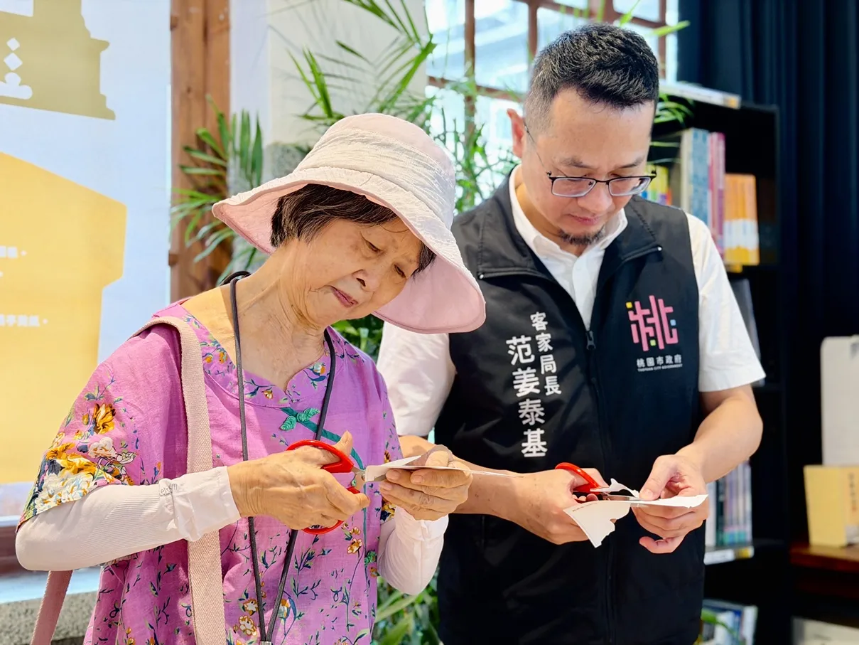 客家局長范姜泰基(右)及李南華女士(左)共同體驗漢聲雜誌曾介紹的剪紙藝術，現場剪出聖蹟亭的剪影。（圖／桃園市政府客家局提供）