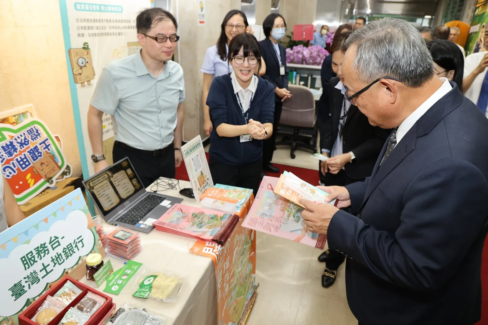 土地銀行80周年，行史檔案展結合公益市集活動，土地銀行總經理張志堅(右)聆聽同仁說明行史中英雙語摺頁設計，接軌國際客群。