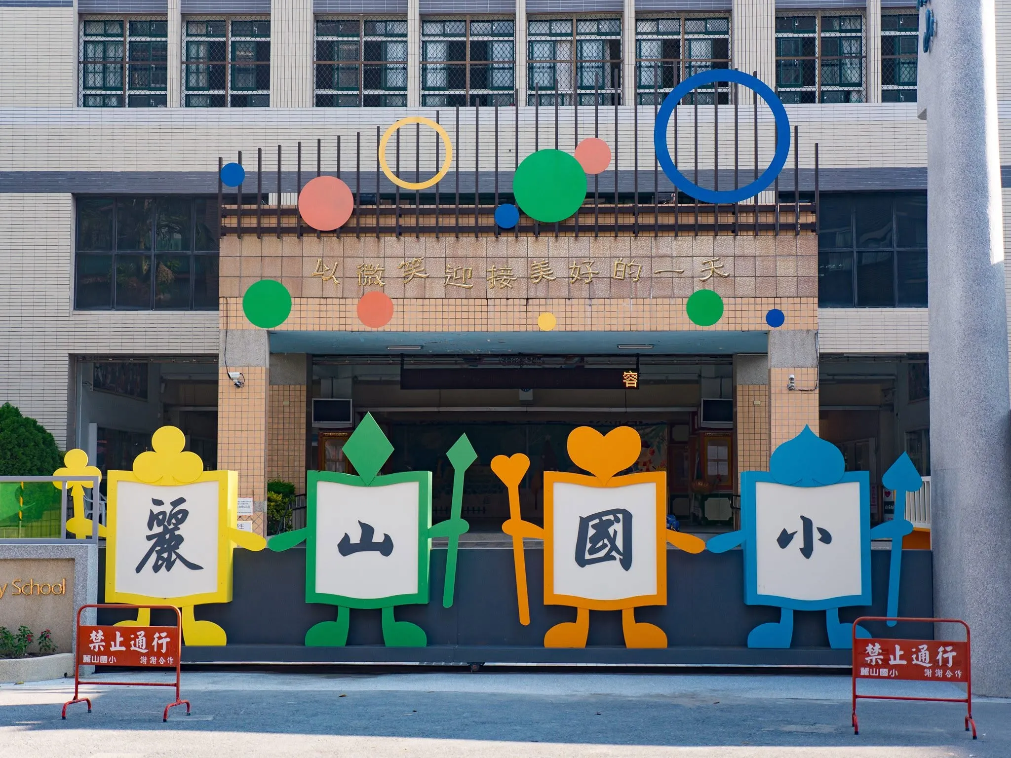 台北市內湖區麗山國小校門口設有彩色裝置藝術設施，以彩色鉛筆造型呈現之學校名稱字樣大看板，校舍牆面還有微笑標語與警告告示。（圖／取自麗山國小）