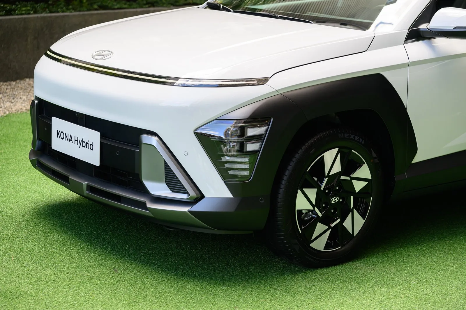 白色 HYUNDAI KONA Hybrid 休旅車頭與單側輪圈特寫，車牌框標示車型名稱，停放於綠色草皮上