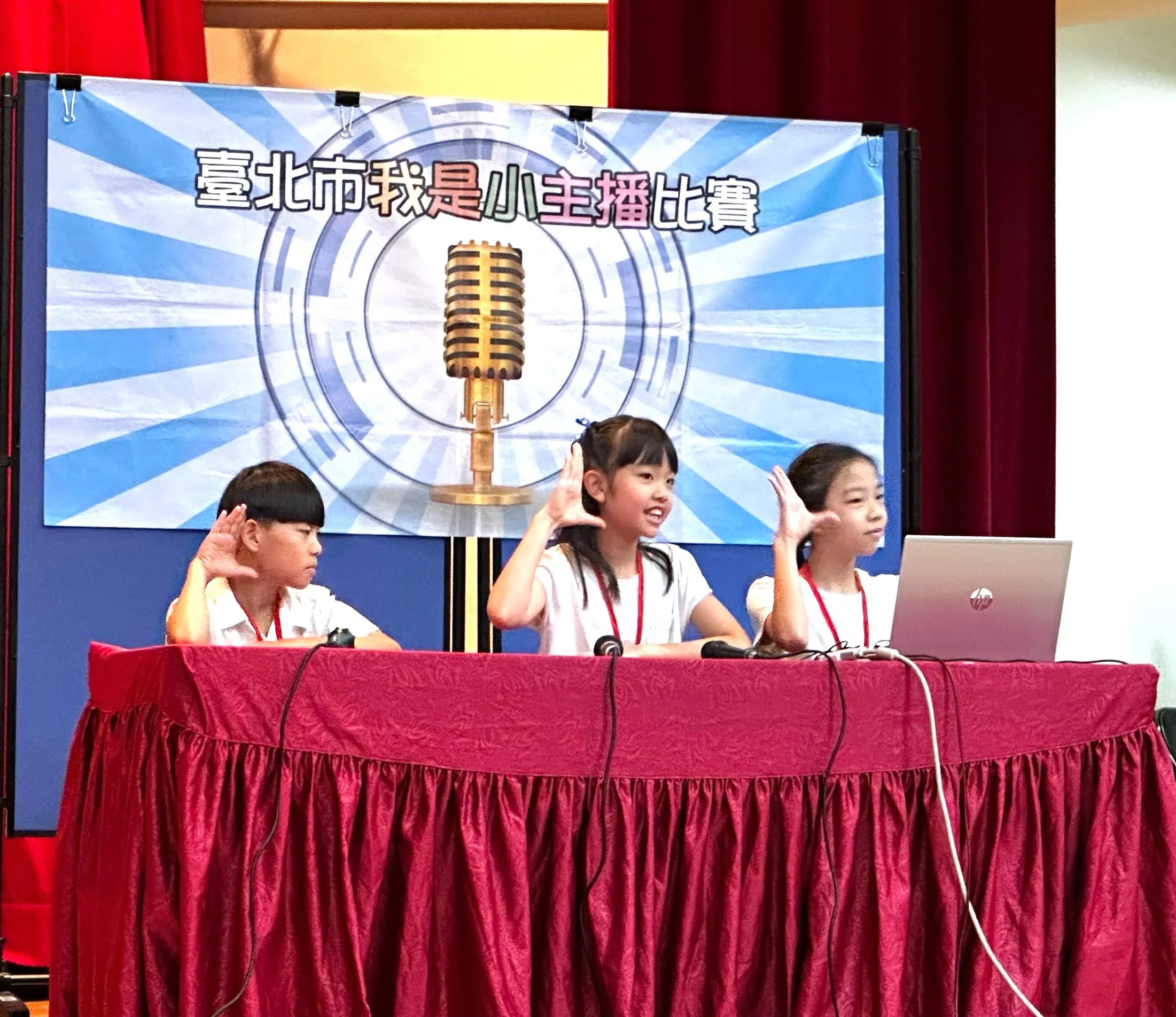 三名參賽學生坐在播報桌前，搭配筆記型電腦、麥克風及手勢呈現內容，在「臺北市我是小主播比賽」舞台上展現口語表達與團隊合作。(圖/台北市教育局提供）