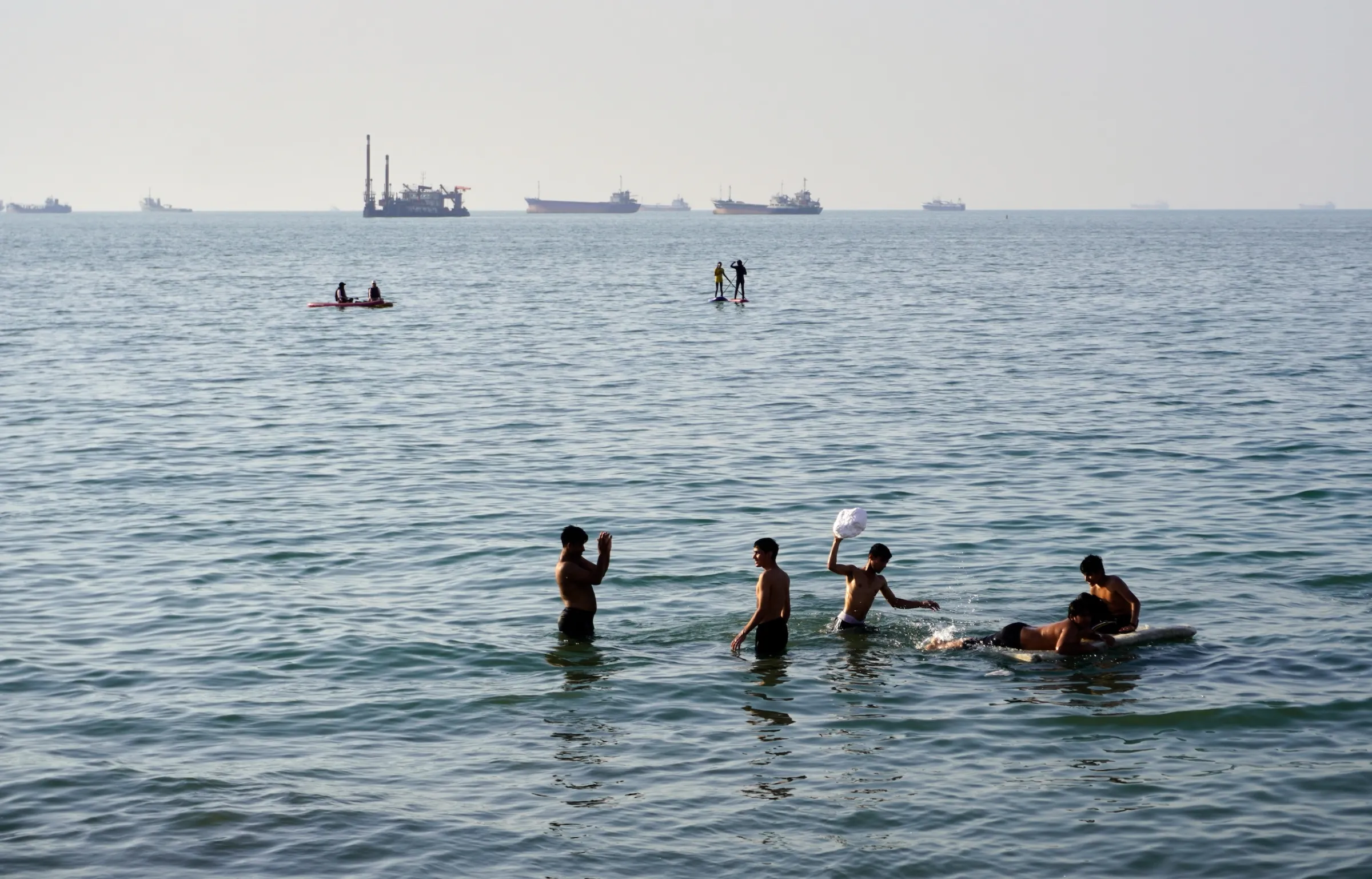 2026年6月10日,伊朗阿巴斯港附近的民眾正在戲水,遠處有停泊在荷莫茲海峽的商船與貨輪。(美聯社)
