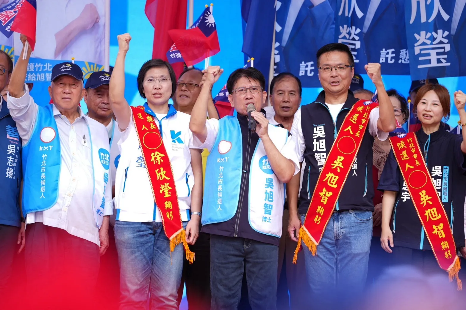 徐欣瑩與吳旭智佩戴候選人綵帶,偕同出席人士在台上舉拳,為縣長及竹北市長選戰凝聚支持。(圖/吳旭智競選總部提供)